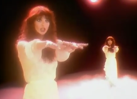 Kate Bush, arms outstretched in a white dress on a black background, shown from two views at once, performing in the music video to Wuthering Heights.