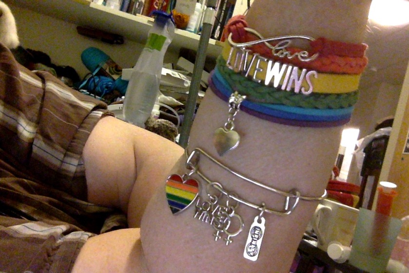 Two Pride bracelets. The first one is a leather rainbow with an infinity sign that says "love", another part that says "LOVE WINS," and a heart dangling off the botom. The second one is a bangle with a rainbow heart, another "LOVE WINS," another infinity, and a double-woman symbol.